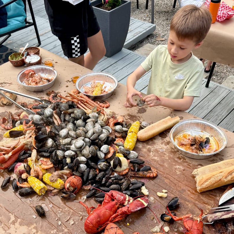A child shucking clams at a seafood boil with lobsters, corn, and mussels on a table.
