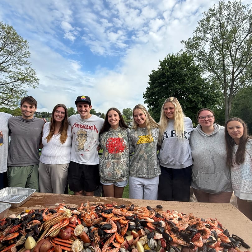 Nine young adults stand behind a table laden with a seafood boil under a cloudy sky.