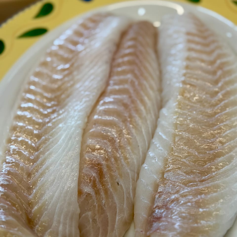 Three raw fish fillets, possibly cod or haddock, on a white plate with a yellow and green rim.